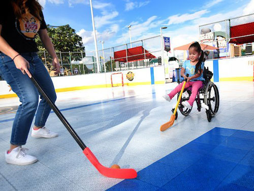 Adult standing with a long hockey stick. Child sitting in wheelchair using a shorter hockey stick.
